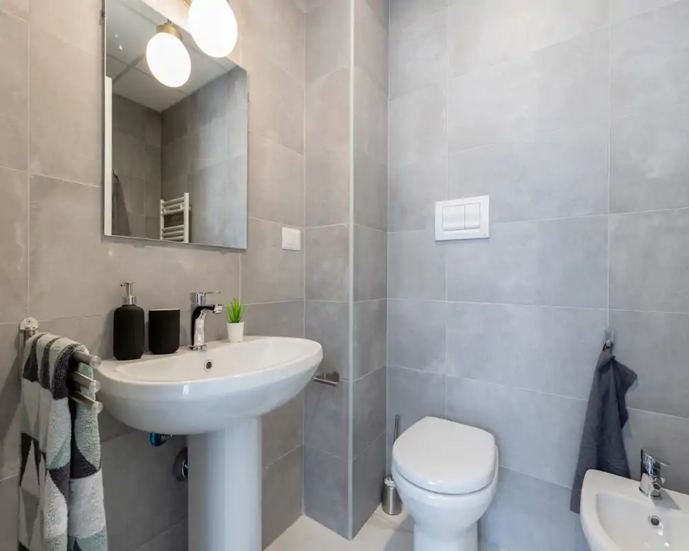 Modern compact bathroom with grey tiled walls, pedestal sink, wall-mounted mirror and toilet; clean, bright and minimalist styling.