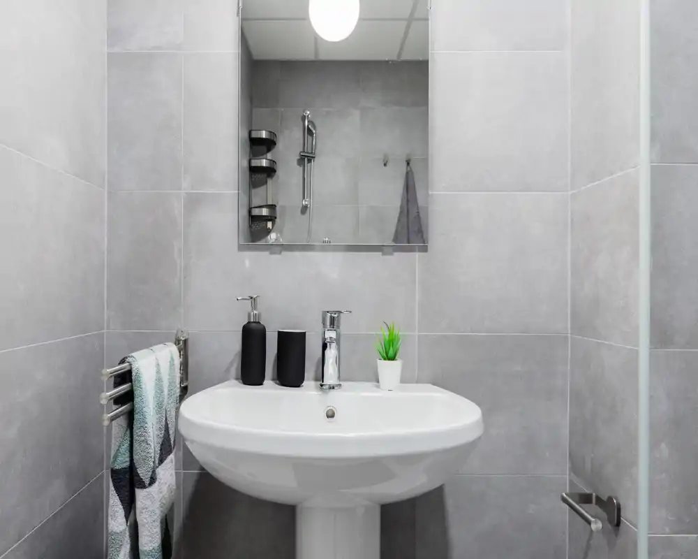 Bright, modern bathroom with a pedestal sink, chrome faucet, mirror and tiled walls — neat styling with soap dispensers and a small plant.