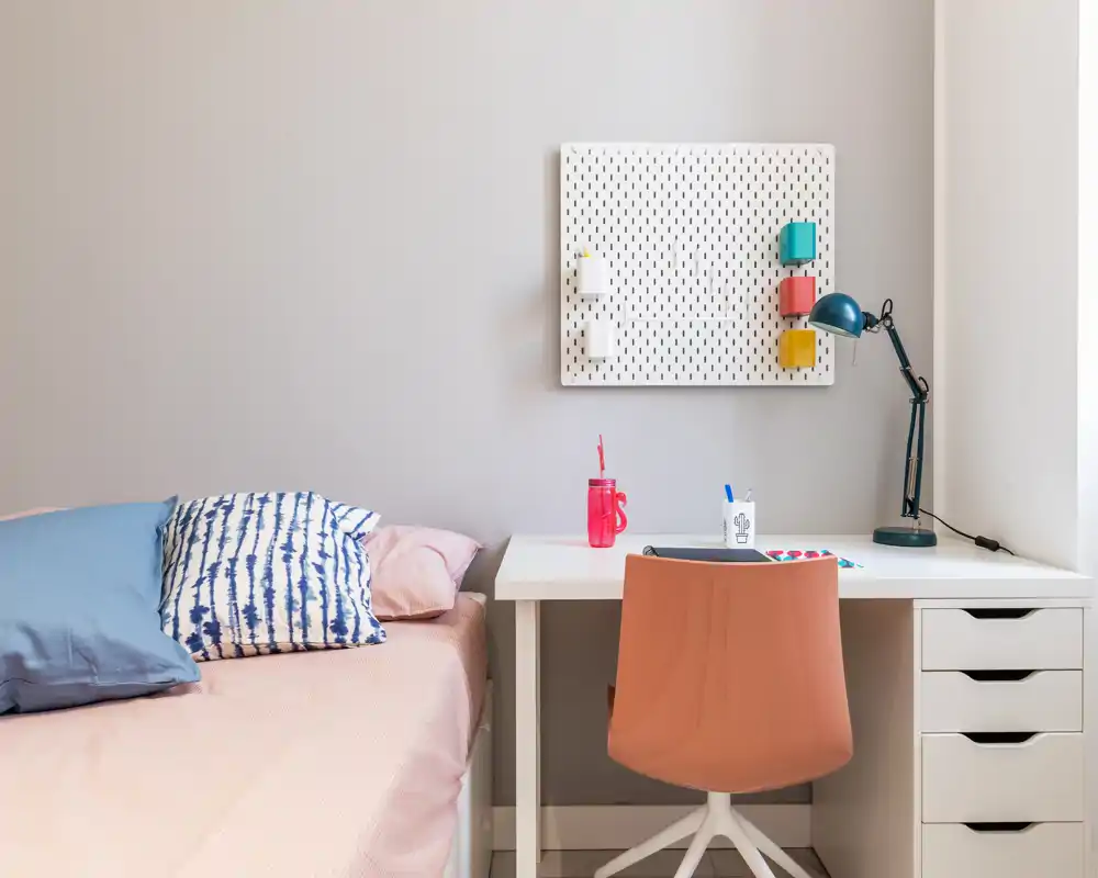 Bright, tidy bedroom corner with a single bed, patterned pillows and a compact white desk with a coral chair — ideal for a student or solo traveler.