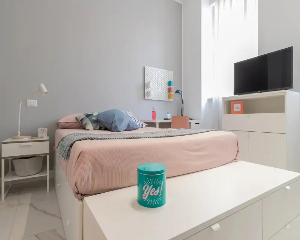 Bright, modern bedroom with a neatly made bed, pastel linens, bedside table and TV cabinet — ideal for a comfortable short-stay.