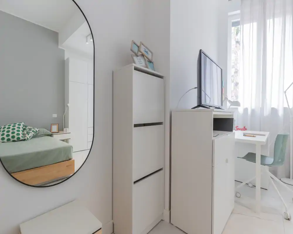 Bright, modern bedroom corner with a bed visible in the mirror, white storage units and a small desk by a sheer-curtained window — minimal, airy styling ideal for rental listings.