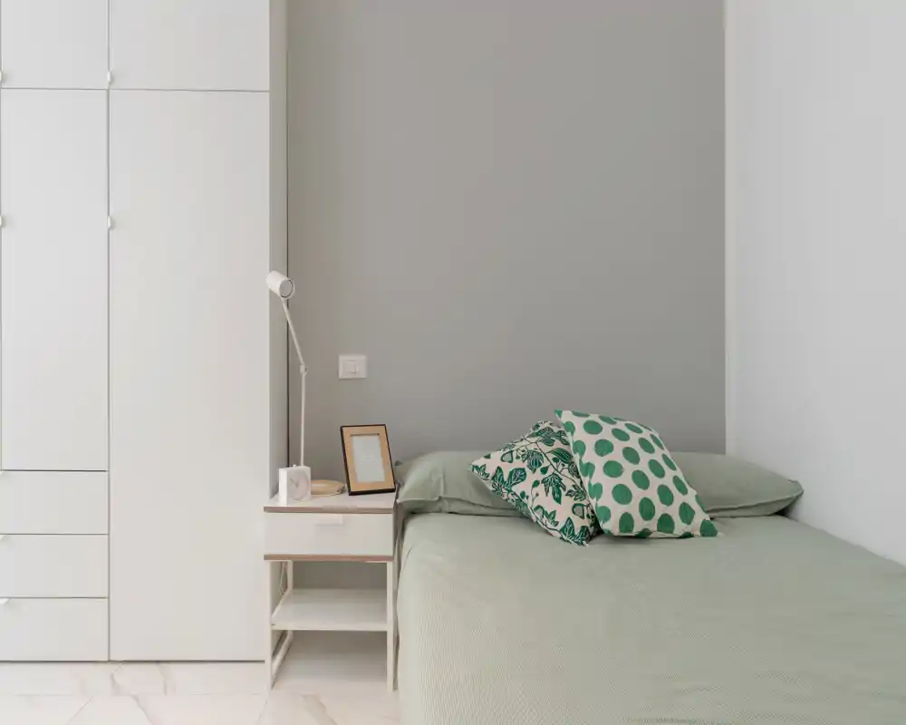 Minimal, modern bedroom with a neatly made double bed, patterned green accent pillows, a bedside table with a lamp and framed photo, and a built-in white wardrobe — bright, calm and uncluttered.