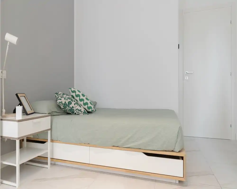 Minimalist bedroom with a low platform bed, soft green bedding and patterned cushions; bright, calm palette and a bedside table with lamp and frame.