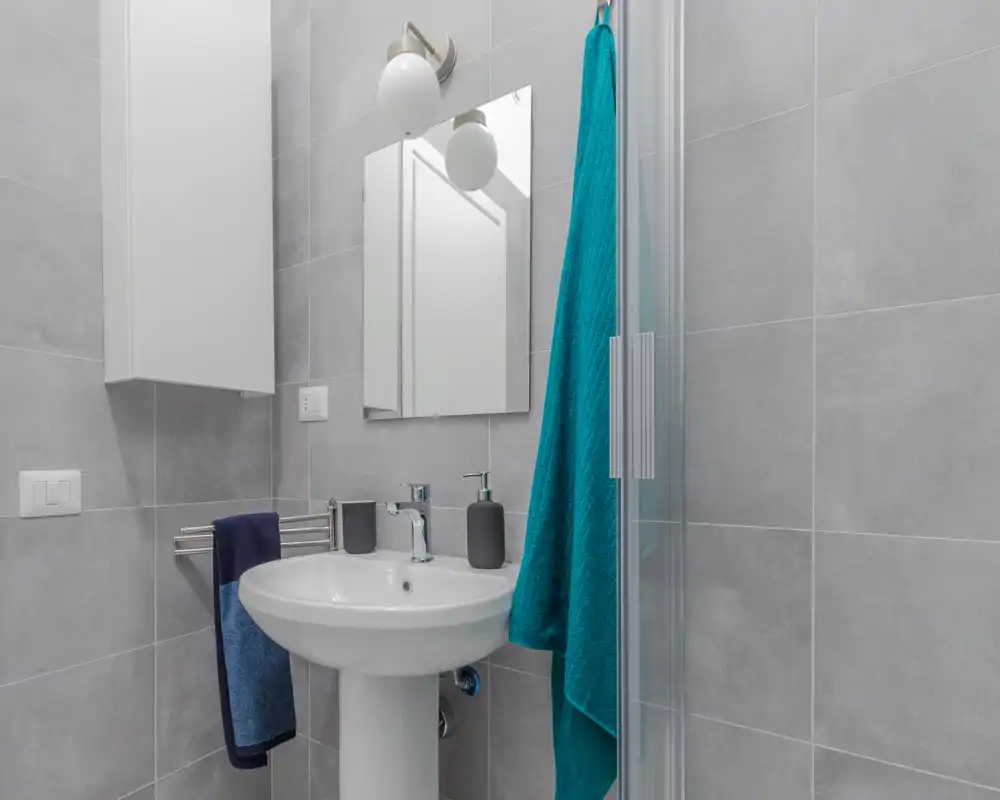 Compact modern bathroom with pedestal sink, mirror, wall cabinet and glass shower door; neutral grey tiles and teal towels add a fresh touch.