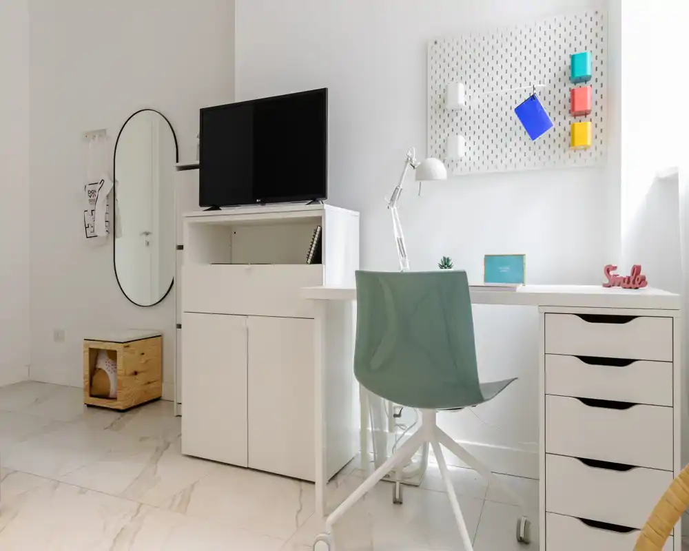 Bright, modern home-office corner with a white desk, storage drawers and a teal swivel chair; tidy, minimal decor including a desk lamp, pegboard and a TV on a cabinet.