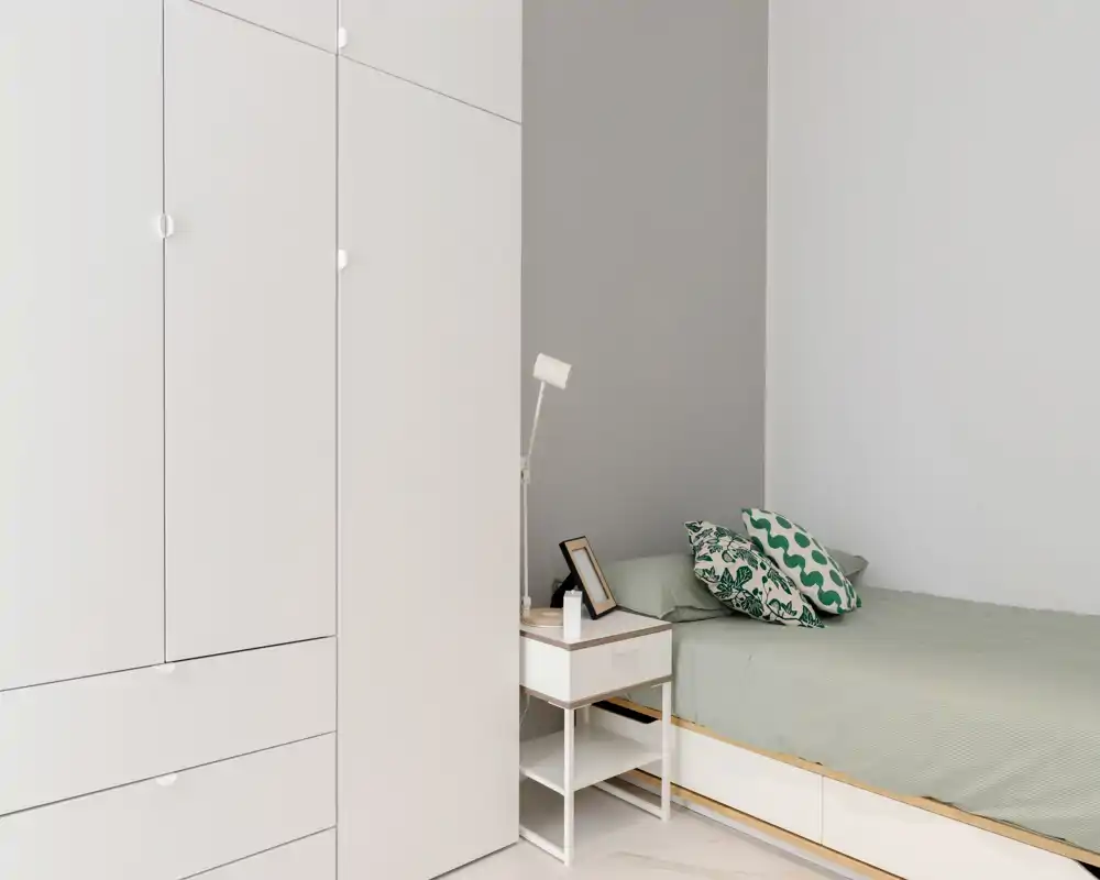 Bright, minimal bedroom with a single bed, patterned cushions and a bedside table with lamp — calm neutral palette ideal for restful stays.