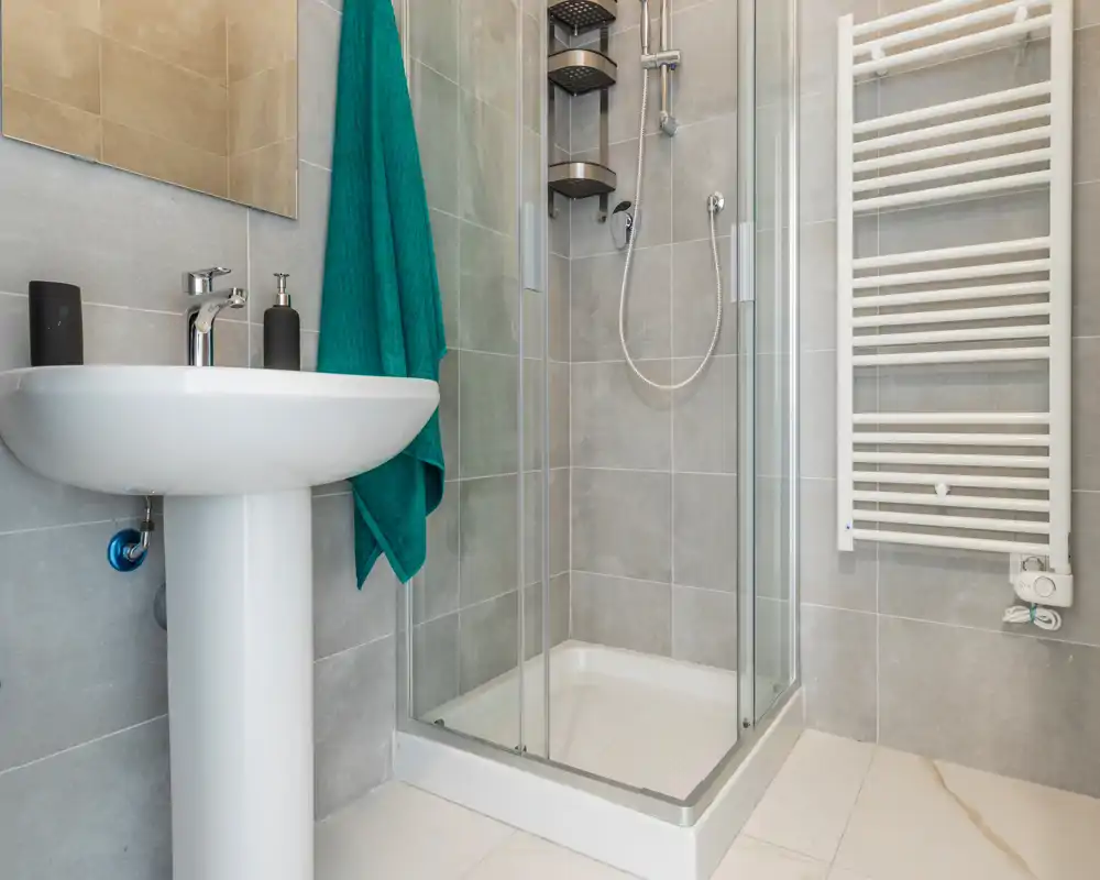 Compact modern bathroom with a glass-enclosed shower, pedestal sink and heated towel rail; neutral grey tiles and a teal towel add a fresh accent.