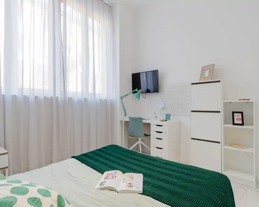 Chambre lumineuse et minimaliste avec lit bien fait, plaid vert et bureau compact sous une télévision murale — idéale pour une personne ou un étudiant.