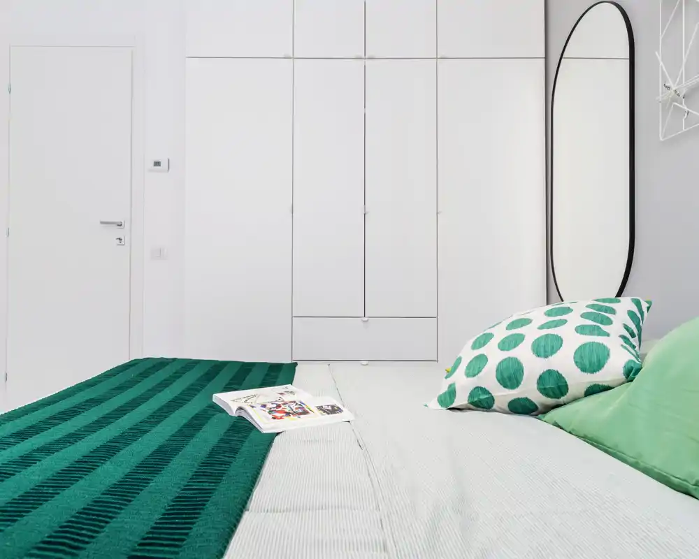 Chambre moderne et lumineuse avec un grand lit au premier plan, un jeté vert et des coussins à motifs, ainsi que des placards intégrés blancs offrant beaucoup de rangement.