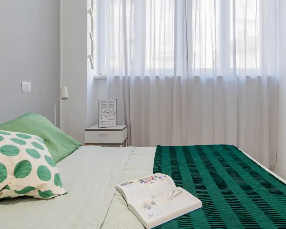Chambre moderne et lumineuse avec un lit bien fait, textiles verts et voilages diffusant la lumière naturelle; table de chevet et coussin décoratif créent une ambiance chaleureuse.