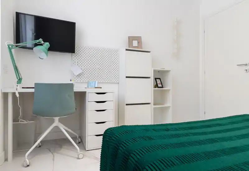Coin de chambre lumineux et moderne avec lit visible recouvert d'une couverture verte texturée, bureau blanc et chaise pivotante, et TV murale — parfait pour une location compacte et bien éclairée.