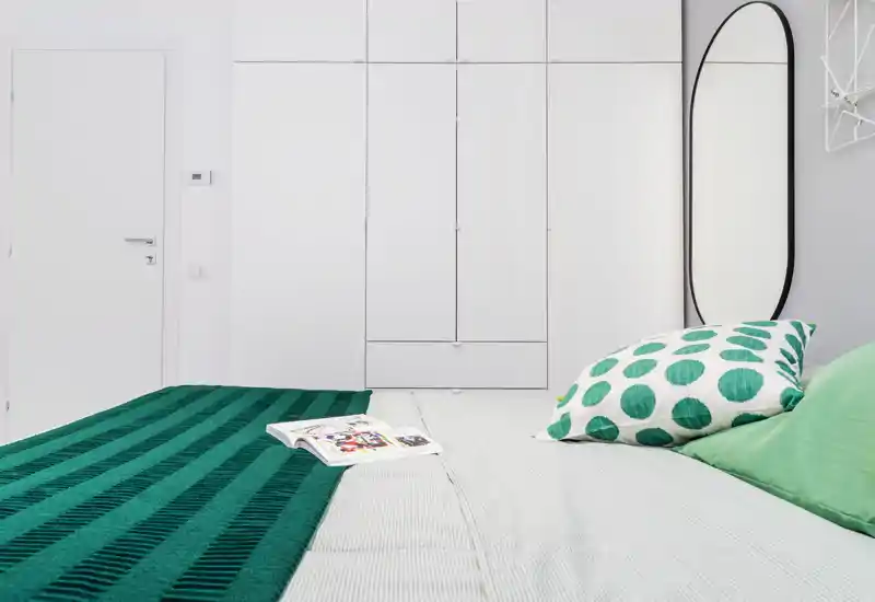 Chambre moderne et lumineuse avec un grand lit au premier plan, un jeté vert et des coussins à motifs, ainsi que des placards intégrés blancs offrant beaucoup de rangement.