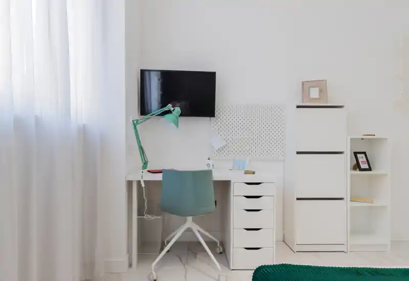 Bright, minimal workspace tucked beside a window — white desk with drawers, a teal task chair and lamp create a clean home-office corner with stylish storage.
