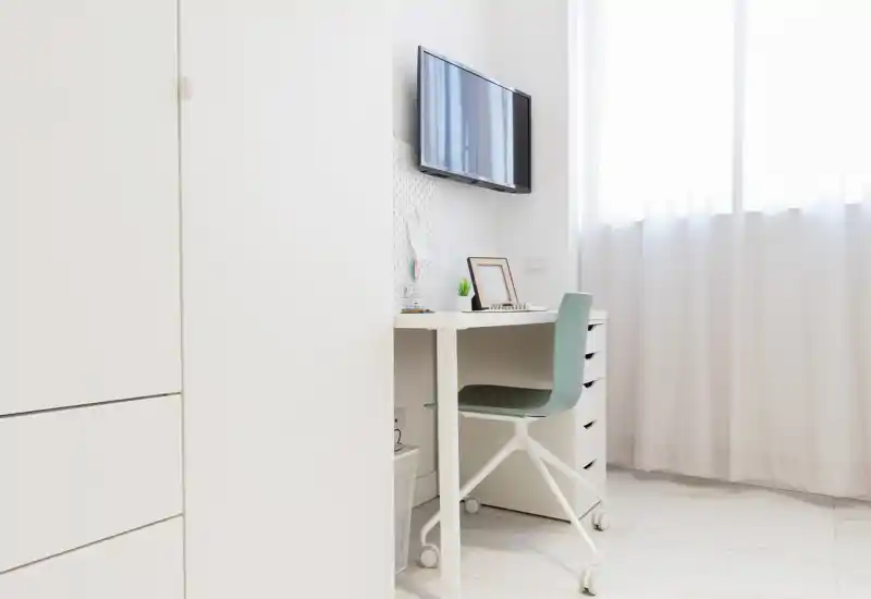 Angolo camera da letto luminoso e minimale con scrivania compatta e sedia su ruote accanto all'armadio; ampie tende leggere lasciano entrare molta luce naturale.