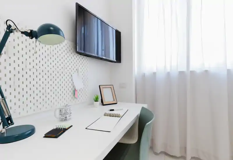 Bright, modern workspace with a clean white desk, adjustable desk lamp, small plant and wall-mounted TV — ideal for focused work or study.