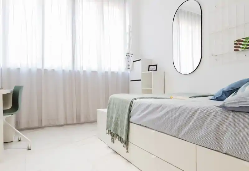 Camera da letto luminosa e moderna con letto ben rifatto, tessili morbidi e una grande finestra che offre molta luce naturale — uno spazio minimal e rilassante ideale per il riposo.