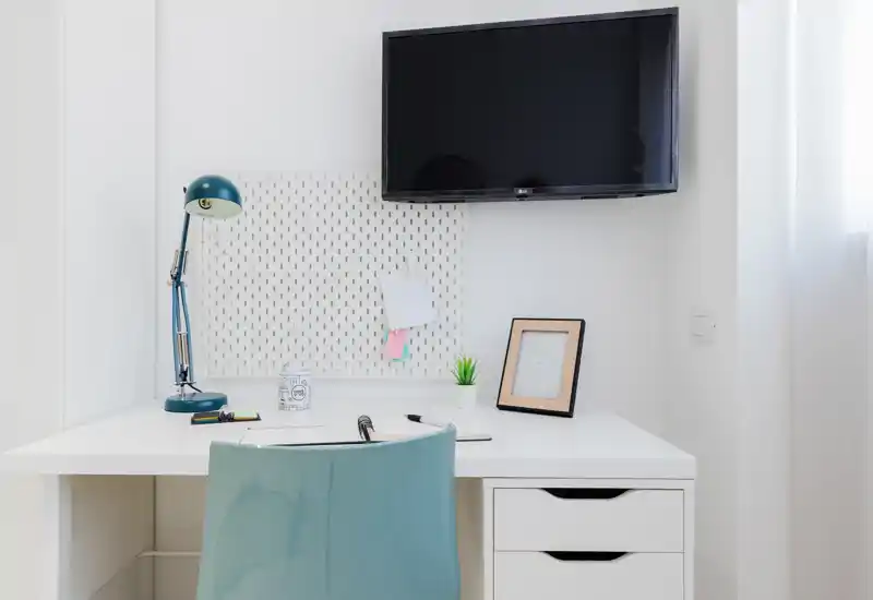 Angolo studio luminoso e minimal con scrivania bianca, lampada e sedia blu, piccola pianta e TV a parete — perfetto per uno spazio di lavoro compatto.