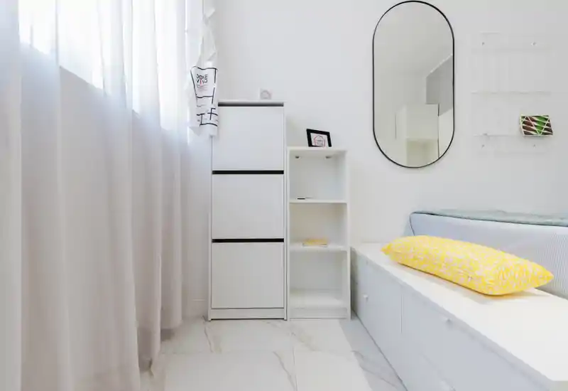 Angolo camera luminoso e minimal con letto basso, cuscino giallo decorativo, mobili contenitori bianchi e uno specchio ovale; stile pulito e arioso adatto a foto per affitto.