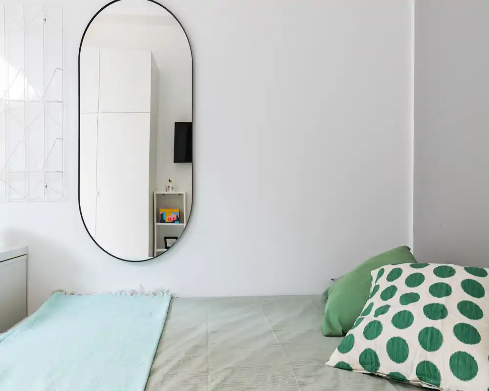 Coin de chambre minimaliste et lumineux avec lit bien fait, coussins verts et grand miroir ovale reflétant une partie de la pièce. Murs blancs et lumière naturelle douce pour une ambiance calme.