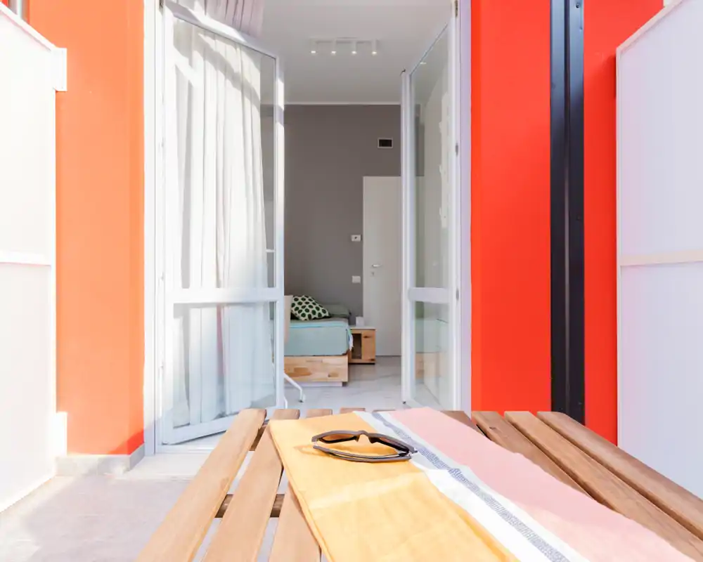 Balcon ensoleillé encadrant une chambre : portes vitrées ouvertes relient une petite terrasse à une chambre soignée avec un lit double visible, mobilier en bois clair et textiles doux.