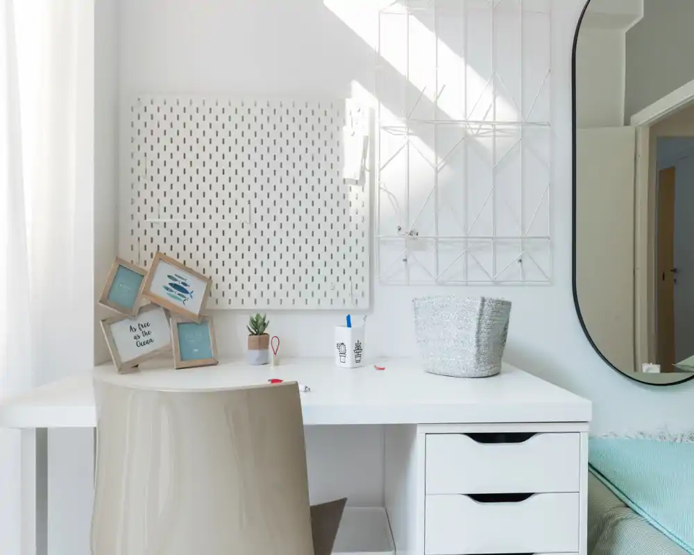Coin chambre lumineux et minimaliste avec un bureau blanc, une chaise simple, des cadres décoratifs et une vue partielle du lit avec un jeté turquoise pâle.