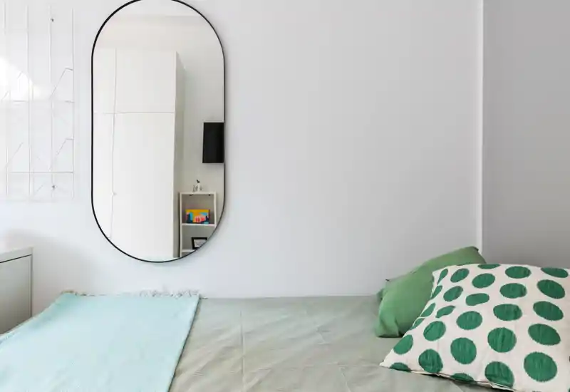 Coin de chambre minimaliste et lumineux avec lit bien fait, coussins verts et grand miroir ovale reflétant une partie de la pièce. Murs blancs et lumière naturelle douce pour une ambiance calme.