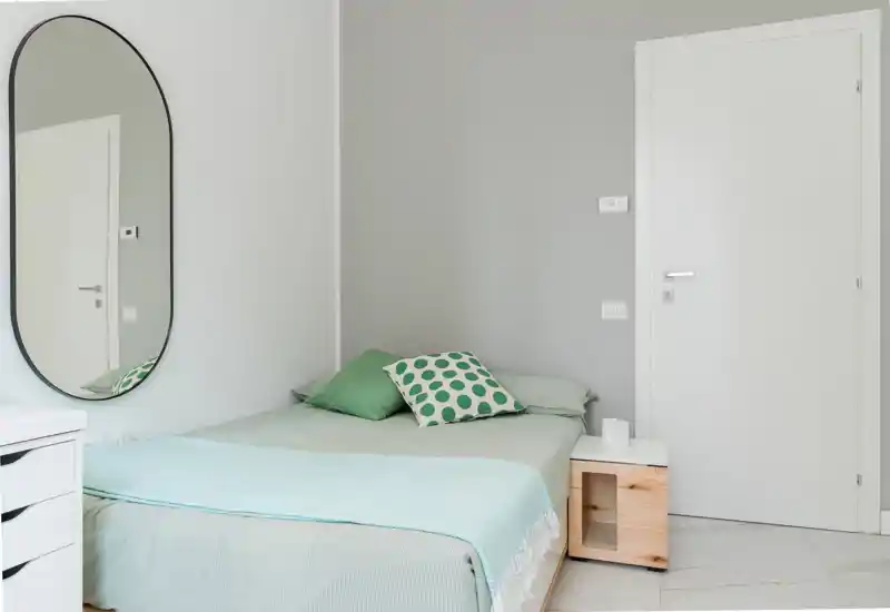 Petite chambre moderne avec lit bien fait, textiles vert pâle, coussin à motifs et miroir ovale mural qui apporte de la luminosité.