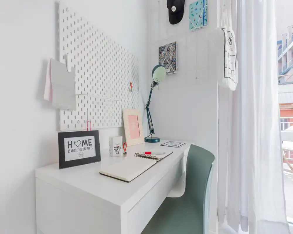 Bright, tidy workspace with a white desk, task lamp and decorative accents next to a sheer curtain letting in natural light.
