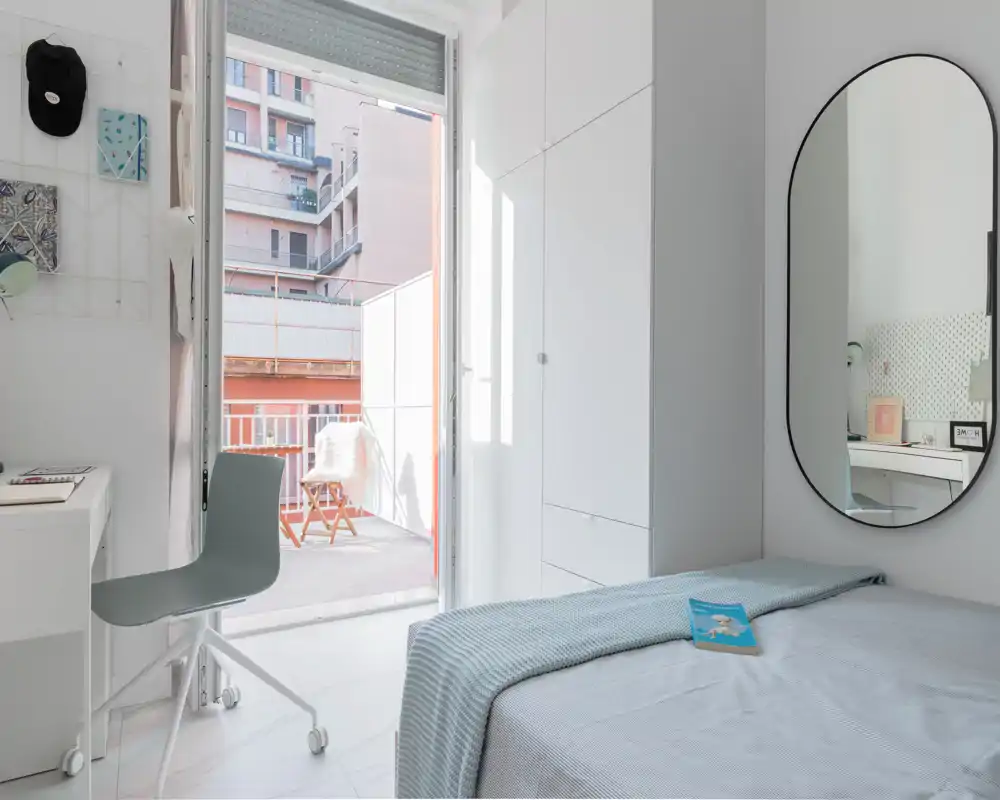 Bright, minimal bedroom with a neatly made single bed, wall mirror and a desk by the window opening onto a small balcony. Natural light fills the room, highlighting clean white cabinetry and soft blue linens.