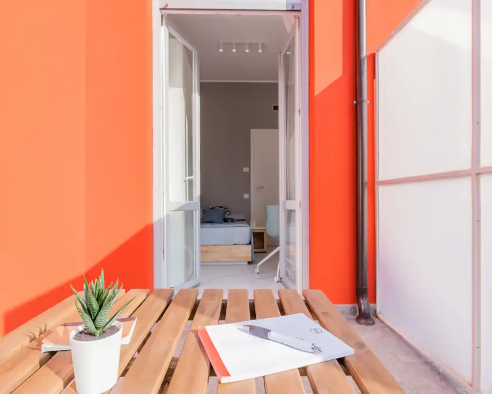 Sunny bedroom seen through open balcony doors, with a wooden bed and bedside table visible; a small outdoor table in foreground creates a welcoming indoor–outdoor feel.