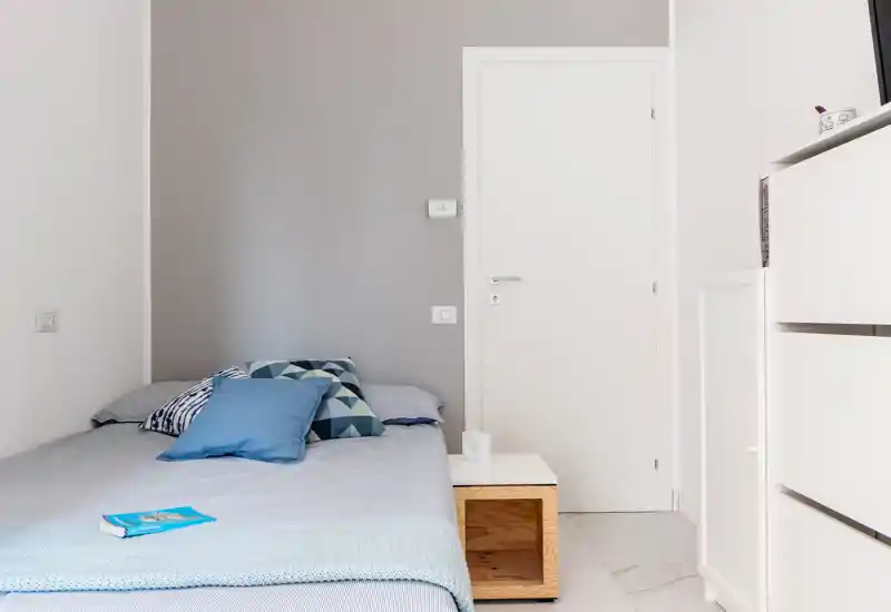 Bright, minimalist bedroom with a neatly made bed, patterned cushions and a small wooden bedside table. Clean, neutral tones and good natural light make this room feel calm and inviting.