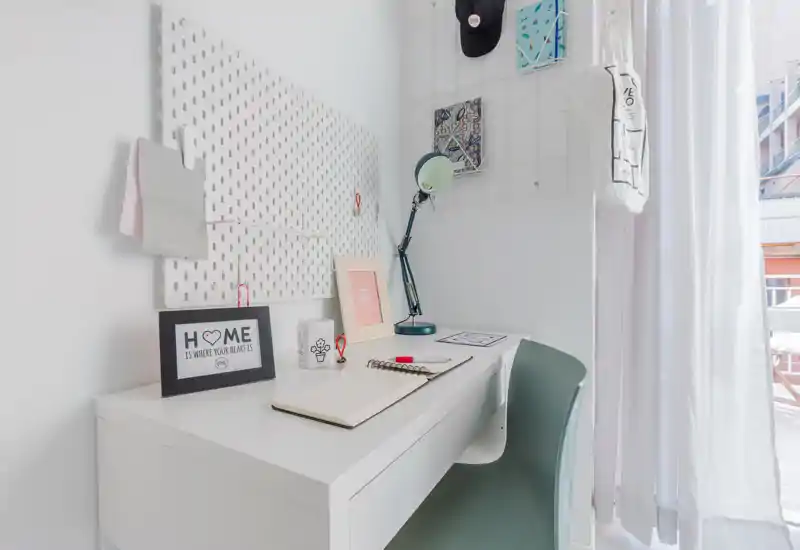 Bright, tidy workspace with a white desk, task lamp and decorative accents next to a sheer curtain letting in natural light.