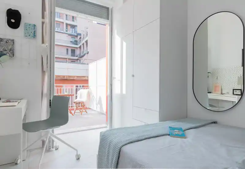 Bright, minimal bedroom with a neatly made single bed, wall mirror and a desk by the window opening onto a small balcony. Natural light fills the room, highlighting clean white cabinetry and soft blue linens.