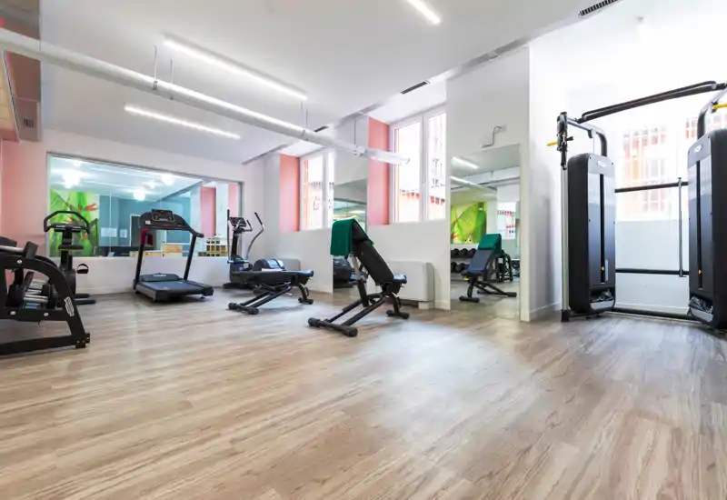 Small private gym/fitness room with cardio machines, adjustable bench and strength station, bright windows and light wood flooring.