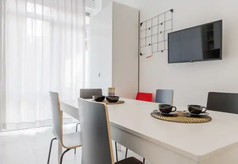 Modern, bright dining area with a white table, six chairs and simple table settings; sheer curtains let in soft natural light for a clean, minimalist look.