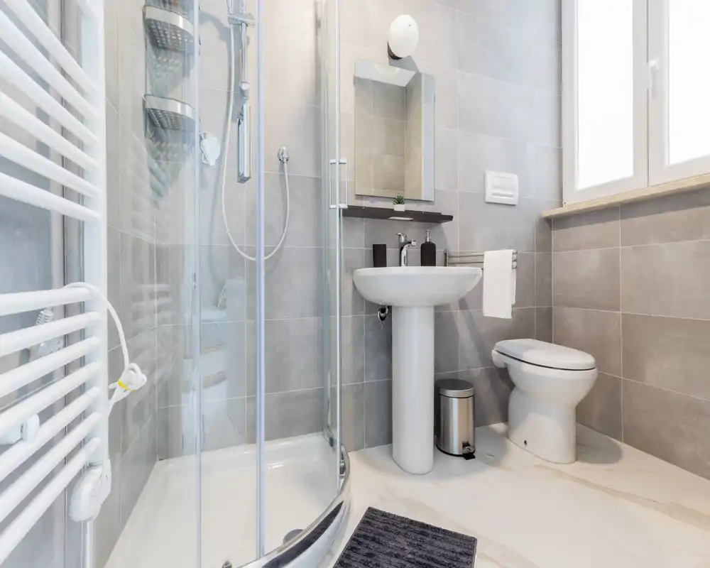 Bright, modern bathroom with glass shower, pedestal sink and toilet; light grey tiles and large window provide a clean, airy feel.