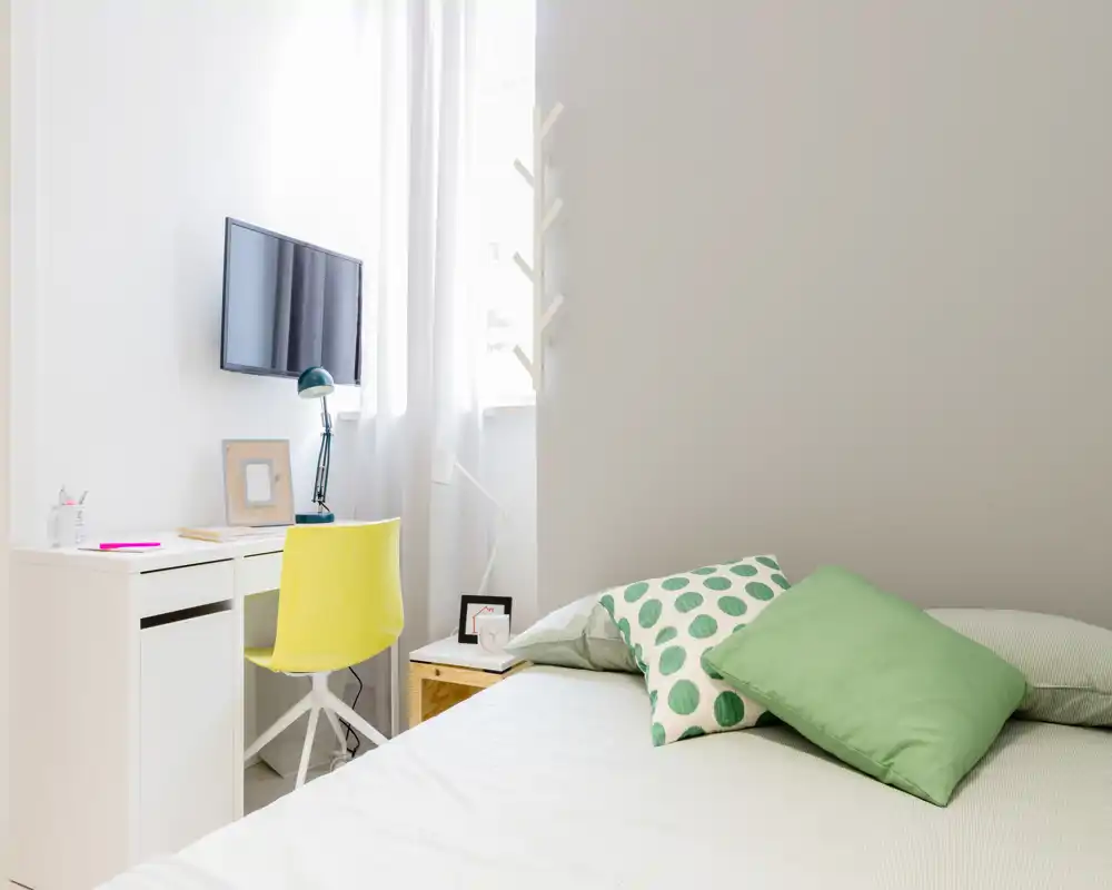 Bright, modern bedroom with a neatly made bed, green accent pillows and a small workspace by the window—ideal for listings highlighting a compact, airy bedroom.