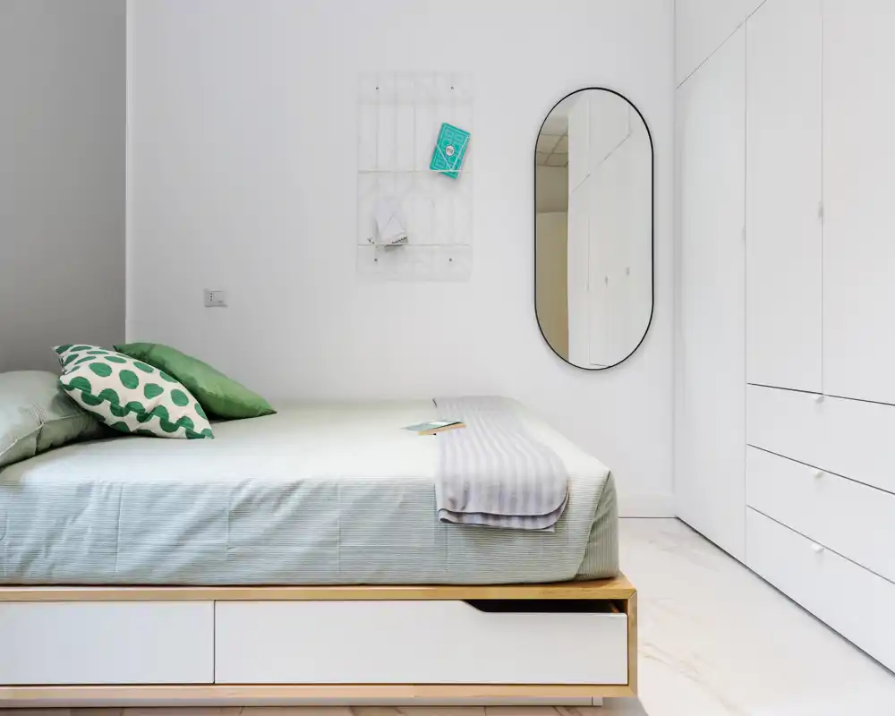 Minimalist bright bedroom with a neatly made bed, green-accent pillows and concealed under-bed storage; white wardrobes and an oval wall mirror create a clean, modern look.