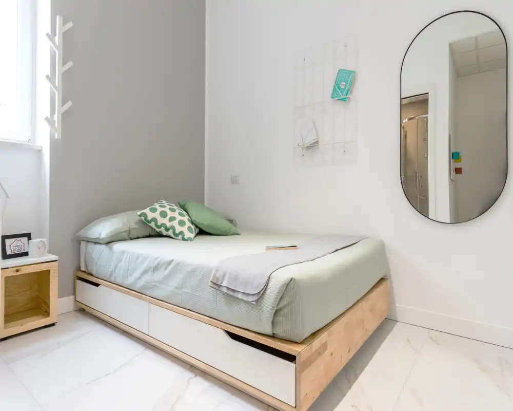 Bright, minimalist bedroom with a platform bed featuring under-bed drawers, soft green bedding and decorative cushions; a large oval mirror adds depth and modern style.
