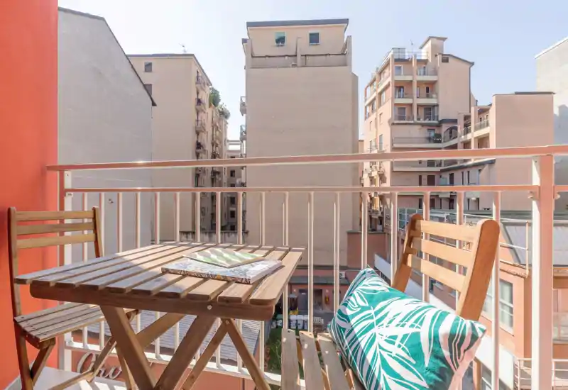 Sunny city balcony with a small wooden table and two chairs, decorative cushion and clear view to neighboring buildings — a compact outdoor spot for morning coffee.