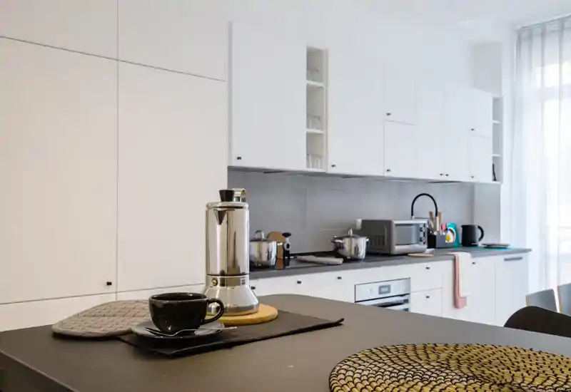 Modern, bright kitchen with white cabinets, built-in oven and countertop appliances; a styled island foreground with a coffee cup and Moka pot creates a welcoming, functional space.