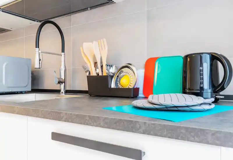 Compact, modern kitchenette counter with a chrome faucet, organized utensils, colorful cutting boards and an electric kettle — bright and tidy.