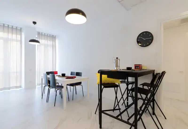 Bright, modern dining area with two tables and multiple chairs, large windows with sheer curtains and contemporary pendant lights create an airy, minimalist atmosphere.
