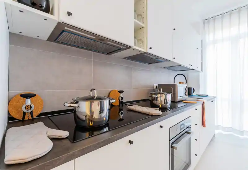 Modern white kitchen with a sleek black countertop, induction cooktop, pots and small appliances; bright natural light through sheer curtains creates a clean, welcoming cooking space.