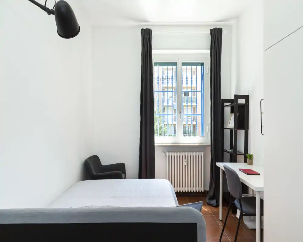 Dormitorio luminoso y minimalista con cama doble, escritorio y butaca junto a la ventana con cortinas oscuras: espacio limpio, moderno y funcional, ideal para una o dos personas.