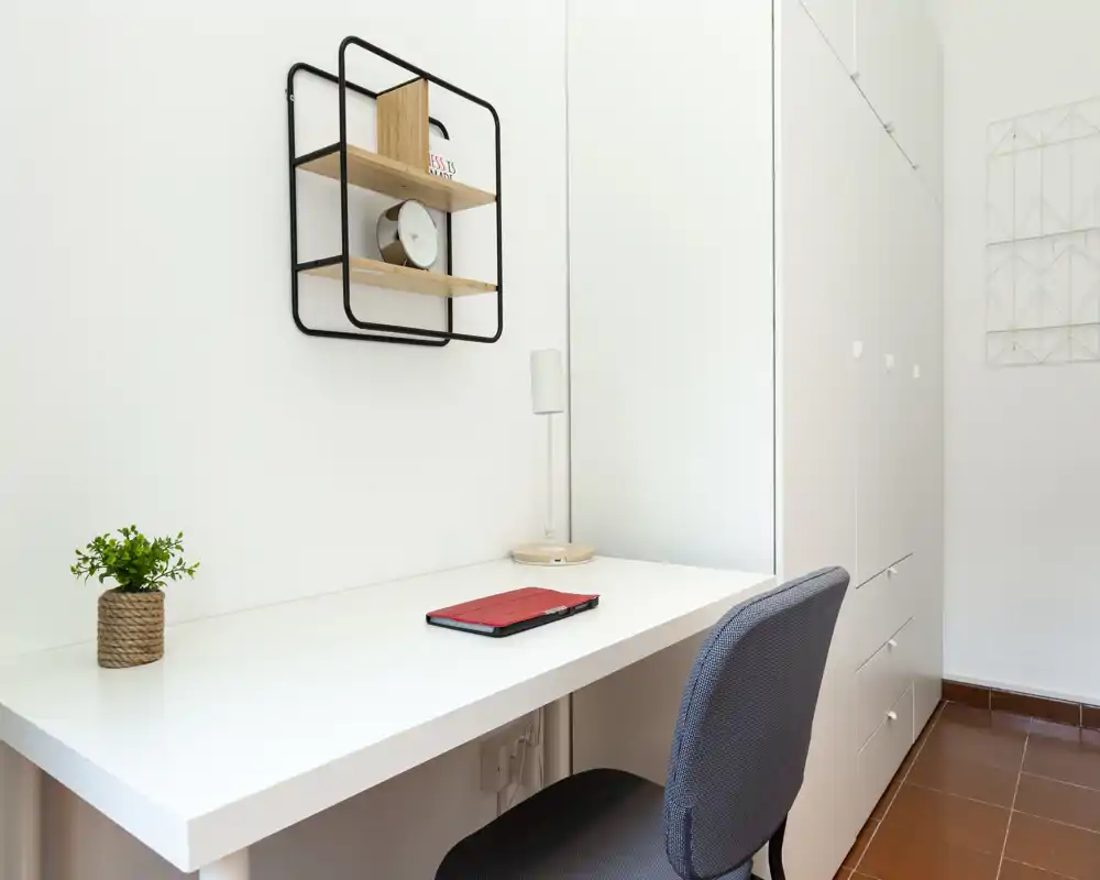 Zona de trabajo luminosa y minimalista con escritorio blanco, planta pequeña, estantería de pared y silla cómoda — ideal para una oficina en casa compacta.