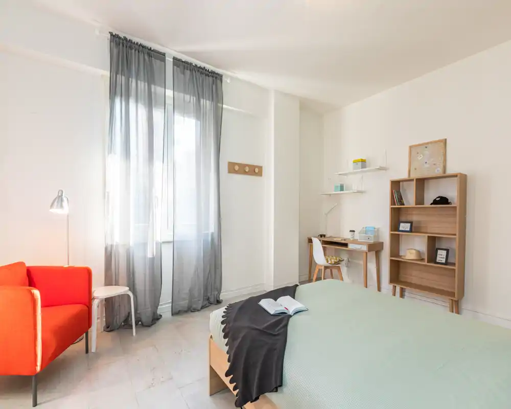 Bright, minimal bedroom with a neatly made double bed, reading throw, desk area and a red accent armchair by the window letting in soft natural light.