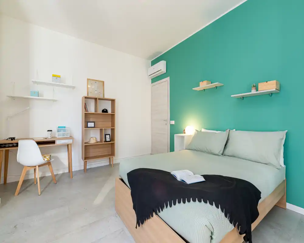 Bright, modern bedroom with a queen bed positioned against a teal accent wall, bedside lamp, simple shelving and a compact workspace — clean lines and soft textiles create a welcoming atmosphere.