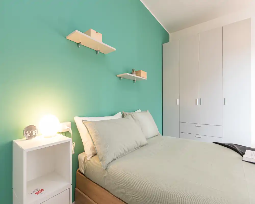 Bright, neatly staged bedroom with a double bed against a mint-green accent wall, bedside table with lamp and a built-in wardrobe providing ample storage.