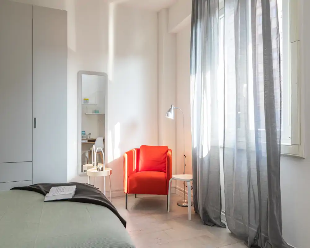 Bright, modern bedroom with a visible bed in the foreground, a red accent armchair by the window and soft natural light creating a warm, inviting atmosphere.
